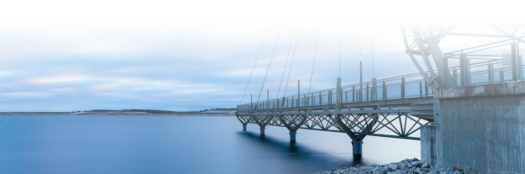 Seebrücke Großräschen - Metallsteg mit Geländer und Seilabspannungen führt von einem Betonsockel über ruhiges Wasser, im Hintergrund eine flache Landschaft unter bewölktem Himmel.