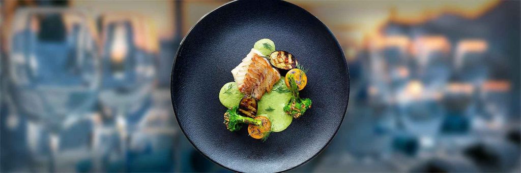 Schwarzer Teller mit einem angerichteten Gericht aus gebratenem Fischfilet, grüner Soße, gegrilltem Gemüse und Brokkoli, von oben fotografiert