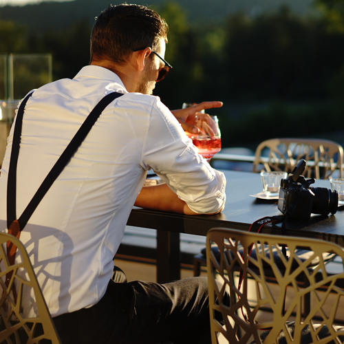 Mann mit Sonnenbrille und Hosenträgern sitzt an einem Tisch im Freien, hält ein Getränk in der Hand, neben ihm liegt eine Kamera.