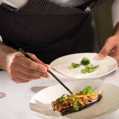 Koch in gestreifter Schürze richtet mit einer Pinzette ein Gericht auf einem weißen Teller an.