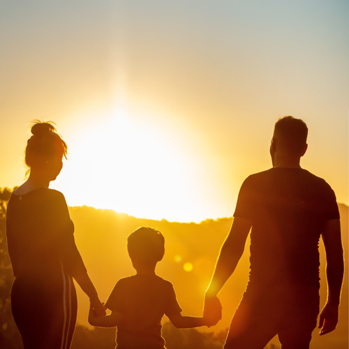 Silhouette von zwei Erwachsenen und einem Kind, die sich an den Händen halten und in den Sonnenuntergang blicken.