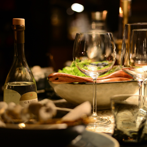Festlich gedeckter Tisch mit Weingläsern, einer Flasche Grappa und einer Schüssel mit Salat im warmen Kerzenlicht.